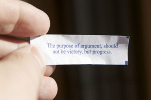 A hand holding a small, typed note that reads, "The purpose of argument should not be victory, but progress."