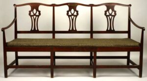 Cherry Chippendale settee; triple-back with serpentine crest rail, scrolled ears, and three pierced scroll splats; rectangular stiles tapering slightly toward top; curved, angled, and slightly outswept arms with scrolled handrest; straight, square legs and rectangular stretchers; inscribed brass plaque at center of crest rail.