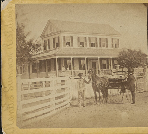 Photograph shows an African American boy holding on to the horse drawn carriage in front of a planter's house. A man prepares to board the carriage.