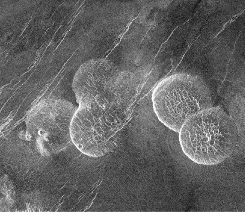 Pancake-shaped volcanoes on Venus. Five of these dome-shaped volcanoes are shown in this image. Three are clustered together in the center left of the photograph, and two others are clustered together in the center right of the photograph.