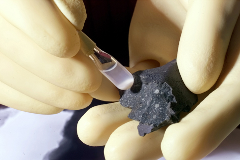 An image of the Murchison meteorite. The meteorite is held in a gloved hand, and another gloved hand holds a test tube containing a small amount of liquid.