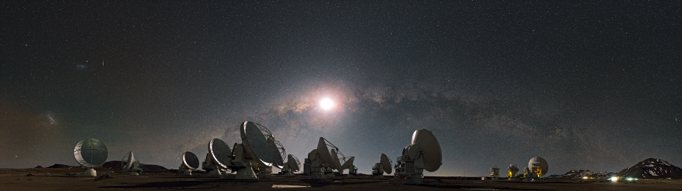 Photograph of the Atacama Large Millimeter Array in Chile, taken at night. Many of the telescopes are seen pointing in various directions, with the Moon and Milky Way prominent in the background sky.