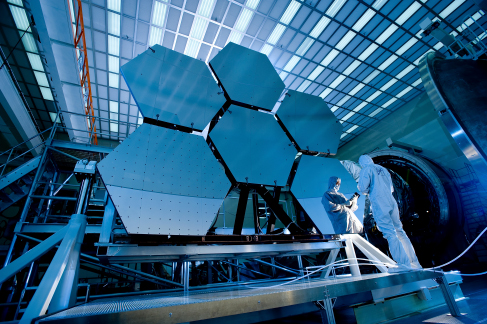 Photograph of the cryogenic mirror testing assembly and 6 of the hexagonal mirror segments of the James Webb Space Telescope. A technician is seen inspecting the right-most mirror segment.