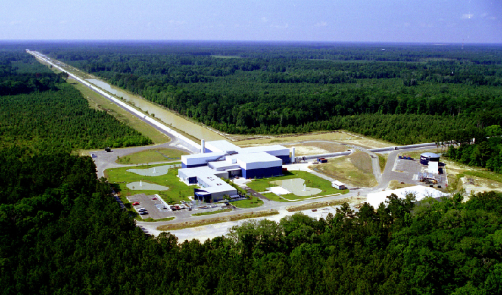 Aerial Photograph of the LIGO Facility. The main building is shown at center, with one of the 4-km long tubes extending toward the horizon at upper left. A portion of the other tube is seen at right.
