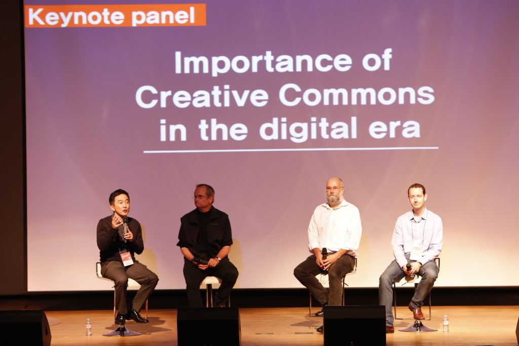 Four men sit on a stage at a conference. Behind them, it says "Importance of Creative Commons in the digital era."