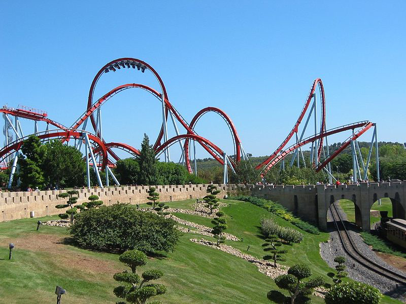 In this figure the Dragon Khan rollercoaster in Spain’s Universal Port Aventura Amusement Park is shown. There are mostly curved paths in the rollercoaster. Near to the rollercoaster there is the track of rollercoaster cart under a bridge. There are some trees near the track.