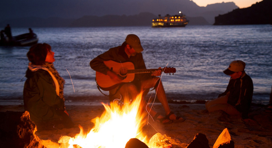 In the figure a couple and their son are sitting alongside a beach in the evening time, around a wood-lit fire. The man is playing a guitar.