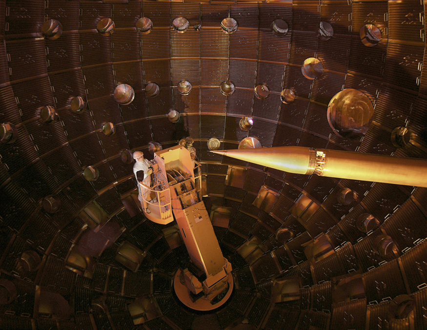 The image shows the inner part of a large shell-like structure where two persons are standing on a boom. The image also shows a sharp pencil shaped structure that serves to hold the fuel pellet at the focus point of all the lasers.