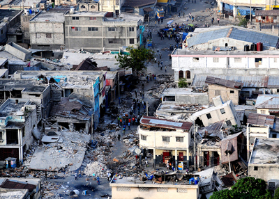 The destruction caused by an earthquake in Port-au-Prince, Haiti. Some buildings are shown on two sides of a street. Two buildings are completely destroyed. Rescue people are seen around.