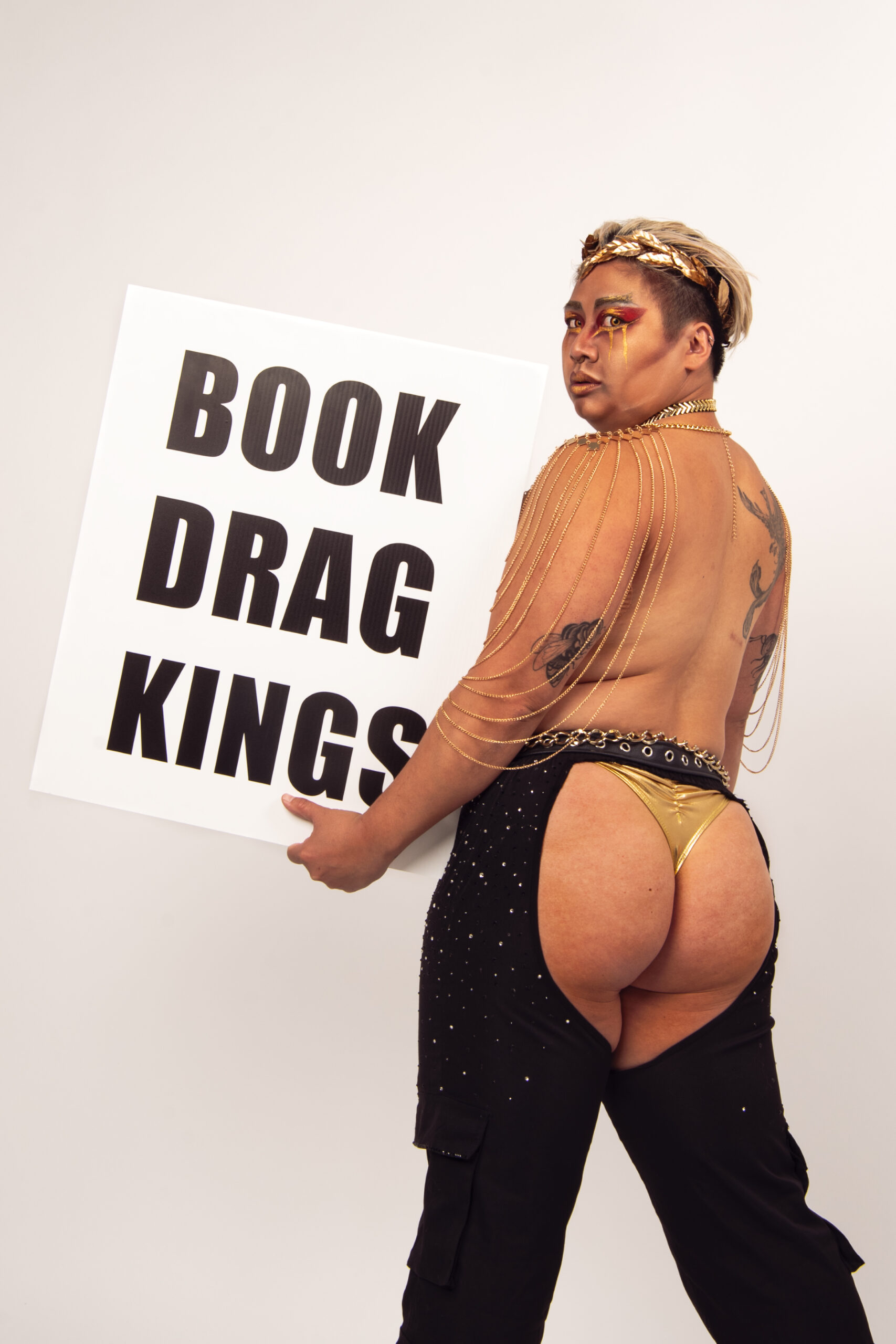 Percy Pegg has his back towards the camera as he looks over his shoulder. He is holding a white sign with bold black text saying book drag kings.