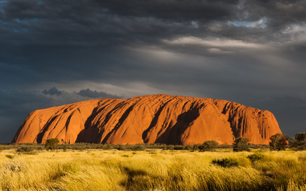 A picture showing Ayers Rock in Australia with a background of dark grey clouds and a forefront of grass