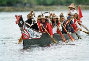 A group of Indigenous people paddling in a canoe that is painted with a traditional design.