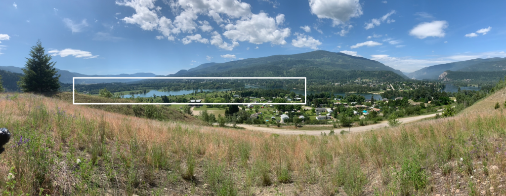 View from a grassy hill looking south over Castlegar Valley from Brilliant Terraces. There is a road at the bottom of the hill with a residential area next to a river with mountains in the background. The area for the sketched map contains the river and anthropogenic development on either side of the river. This particular area is identified on the image with a rectangle.