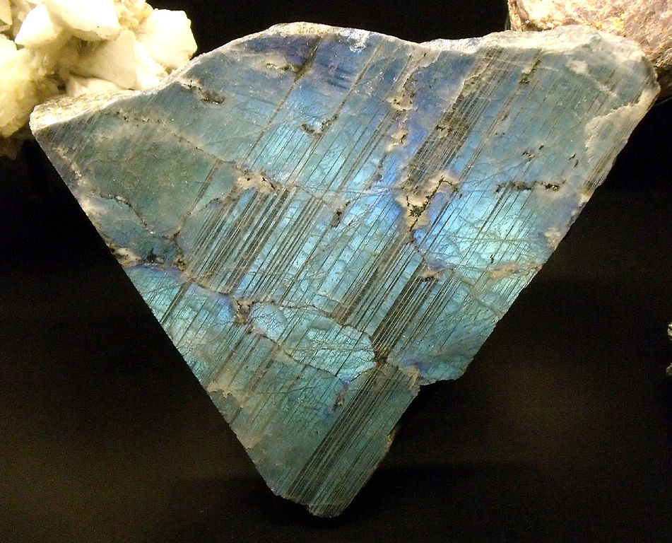 Triangular fragment of a mineral with a surface covered in thin parallel lines. The mineral appears to glow blue and purple.