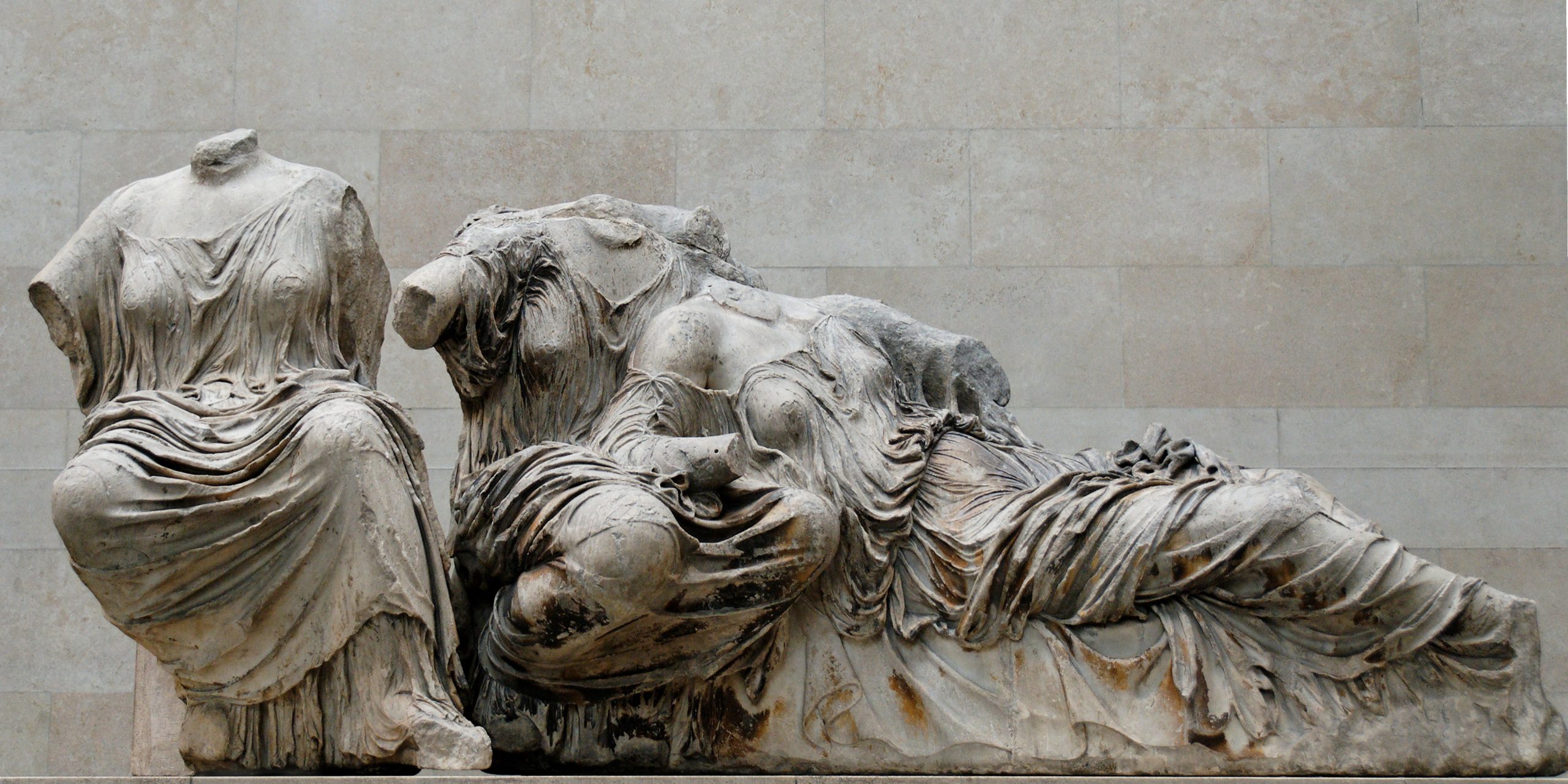 Aphrodite reclining with her head and shoulders on the lap of Dione. To their left sits Hestia. All three are dressed in sheer robes that cling to their forms. The figures are missing their heads and arms.