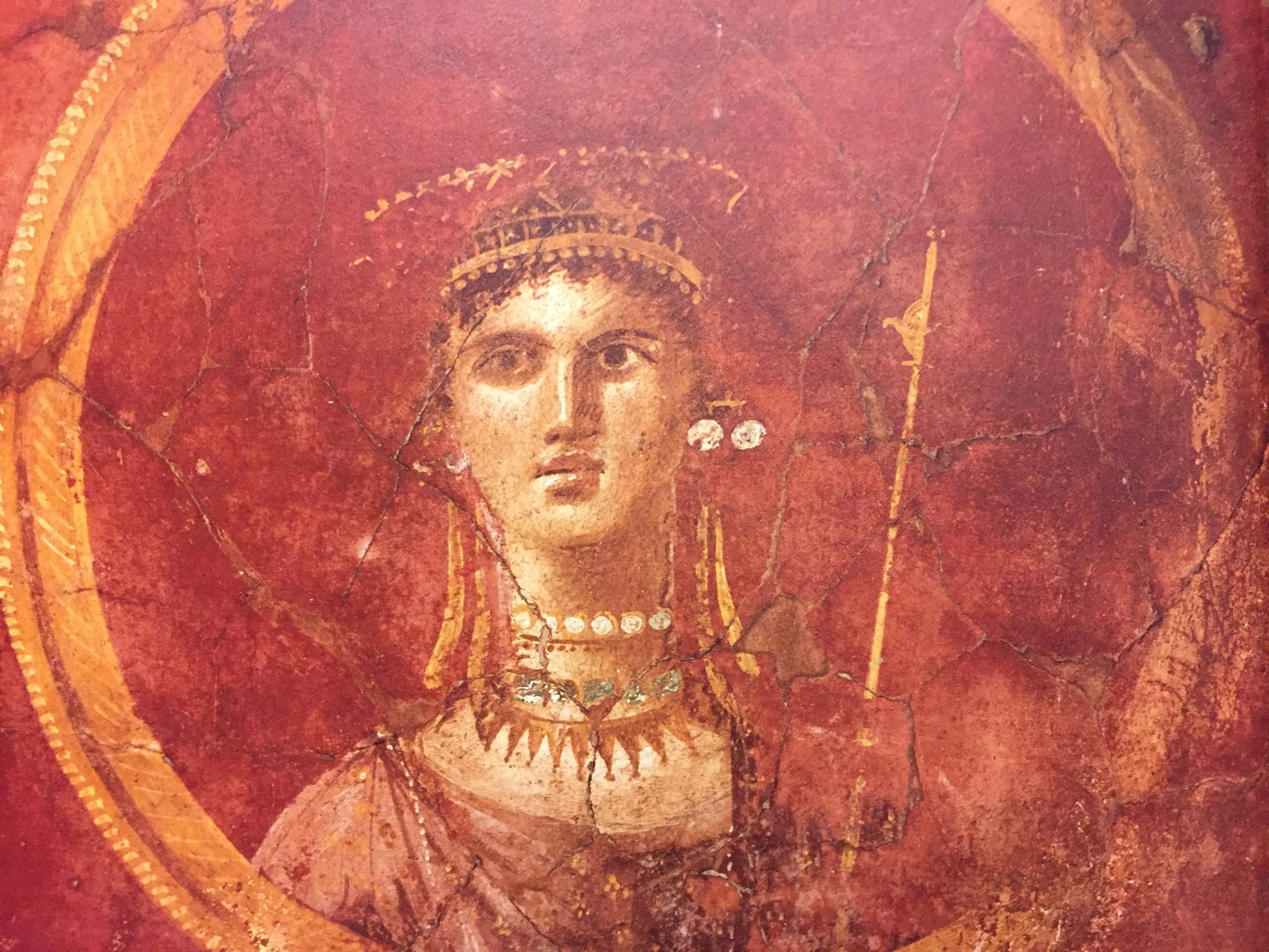 Head and shoulders of Venus wearing a large golden crown and a jeweled necklace. She holds a golden sceptre. Her dress, and the background of the fresco, are in shades of pink and red.