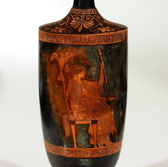 Hera is seated on a throne, dressed in a chiton, holding a staff in one hand and a bowl in the other. There is a falcon perched on the back of the throne.