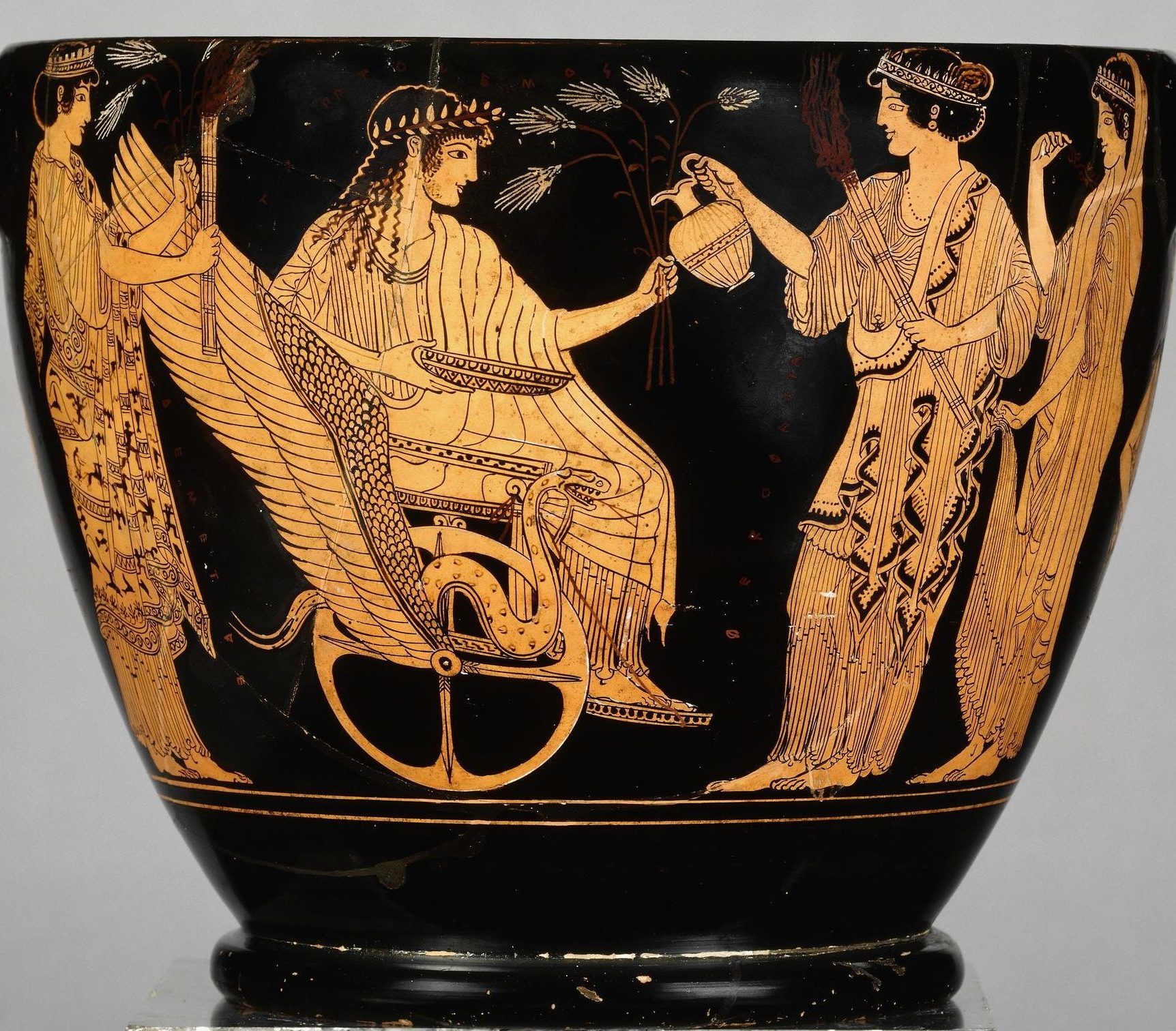 Triptolemos seated in a winged chariot holding sheaves of wheat and a bowl. Persephone, holding a torch, pours from an oinochoe jug into the bowl. Demeter stands behind Triptolemos holding a torch and sheaves of wheat. Eleusis stands behind Demeter.