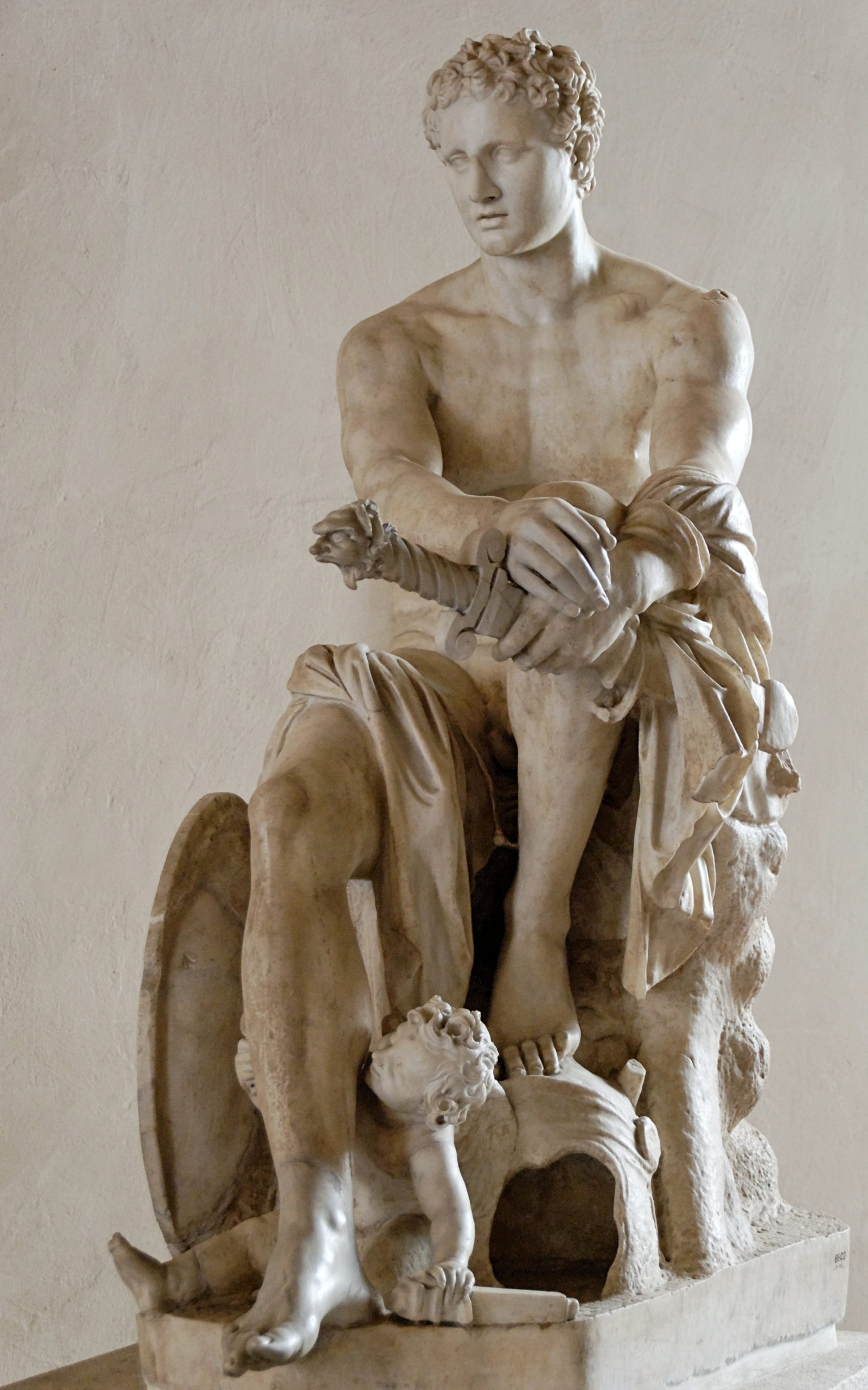 Ares seated with one foot resting on his helm. He is nude save a draped cloth, and is holding a sword. At his feet are a cupid, and Ares' shield.