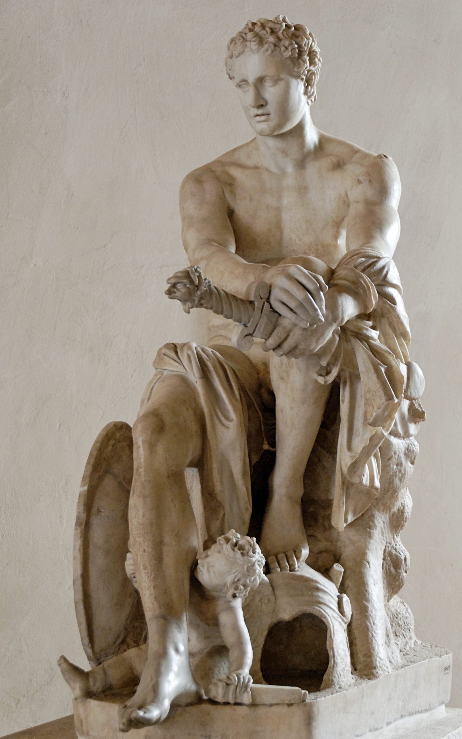 Ares seated with one foot resting on his helm. He is nude save a draped cloth, and is holding a sword. At his feet are a cupid, and Ares&#039; shield.