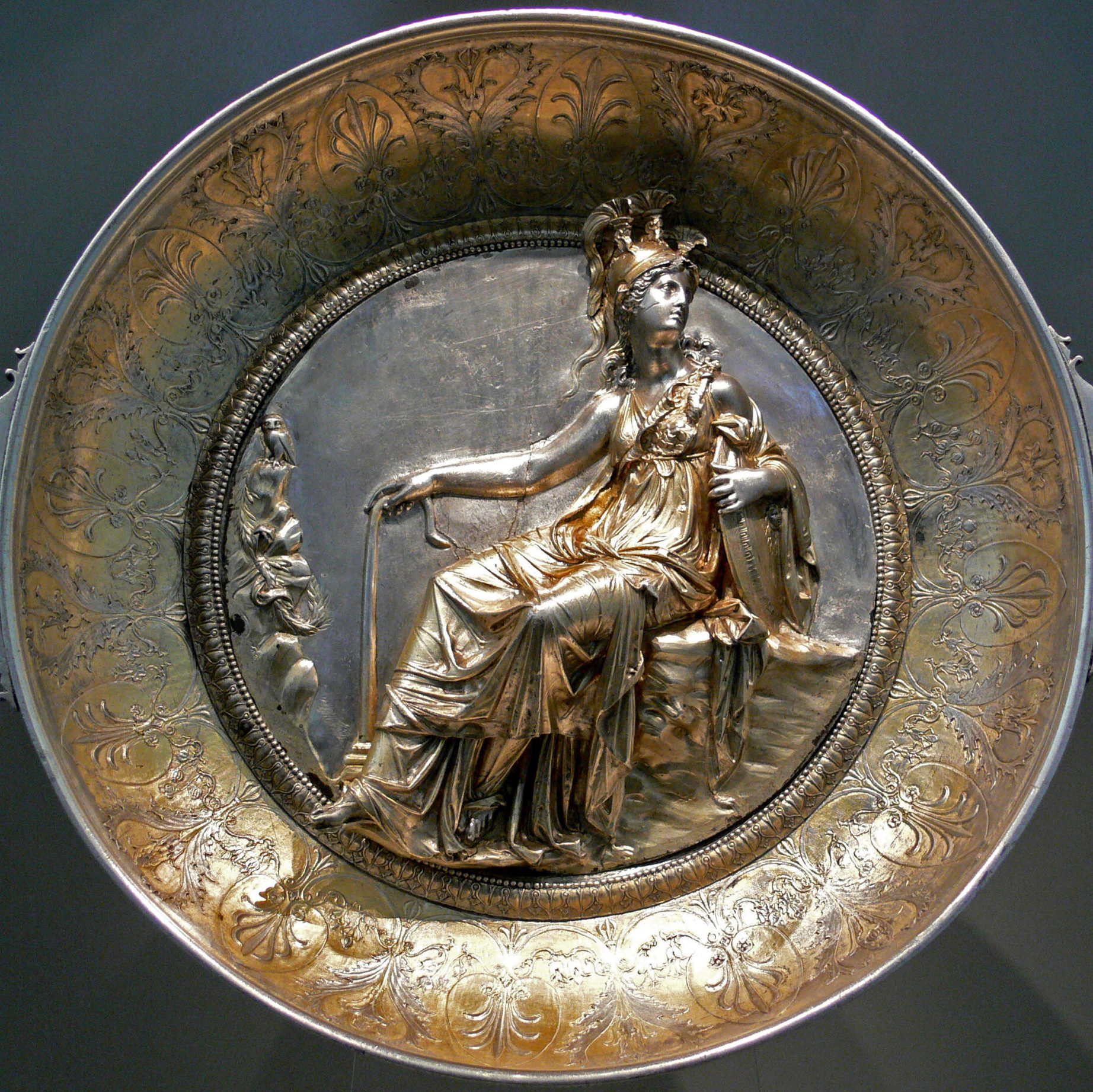 Athena, draped and seated. He is holding a staff in one hand a shield in the other, and has as plumed helm on her head.