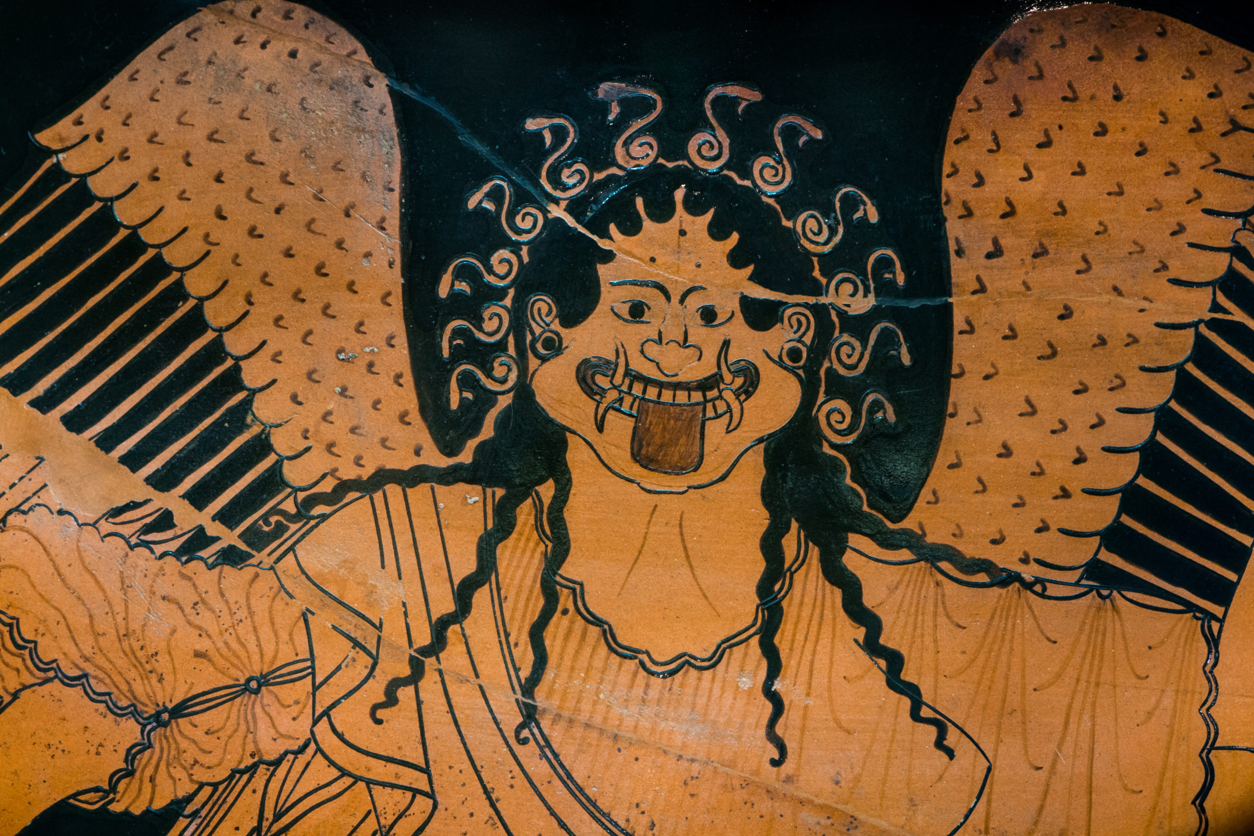 Head and torso of a grinning woman with wings, snakes in her hair, and tusks.