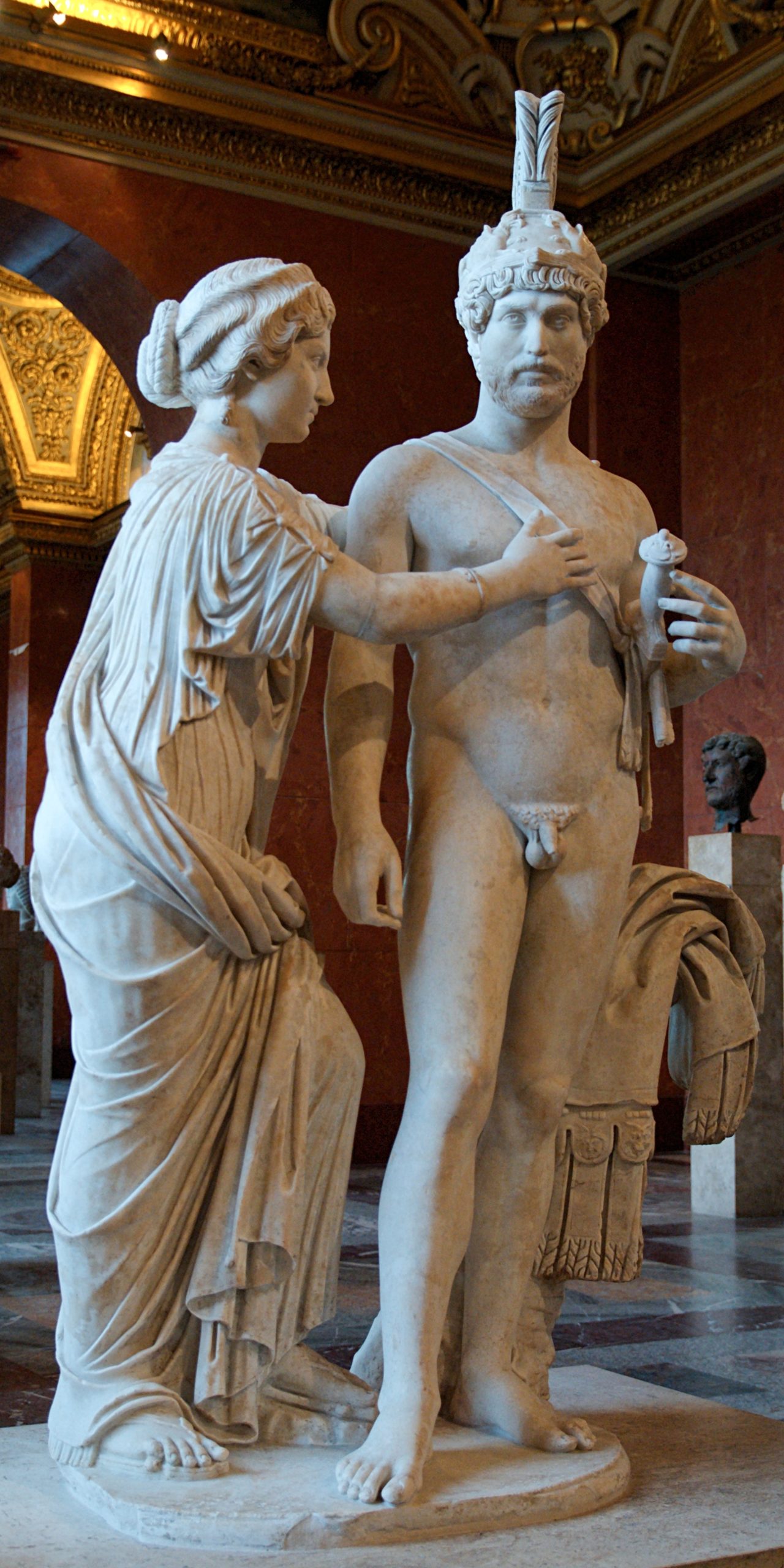 A robed woman stands with her arms around the nude figure of Hadrian. Hadrian is posed like Mars, with a sword at his side and a helm.