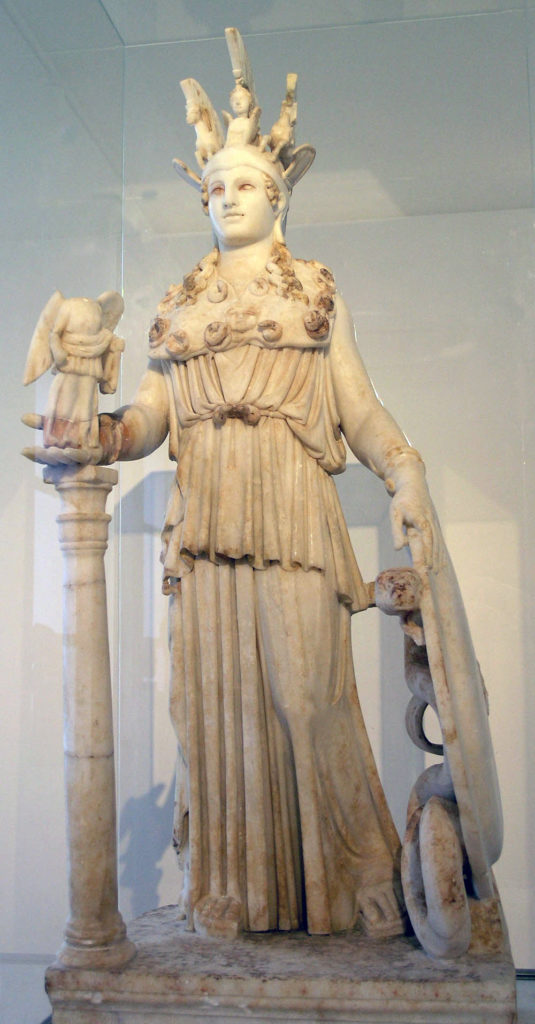 Athena standing. She is wearing an elaborate headdress decorated with figures of horses, and is draped in a peplos. In her right hand she holds a statue of the winged goddess Nike, and in her left she holds a shield.