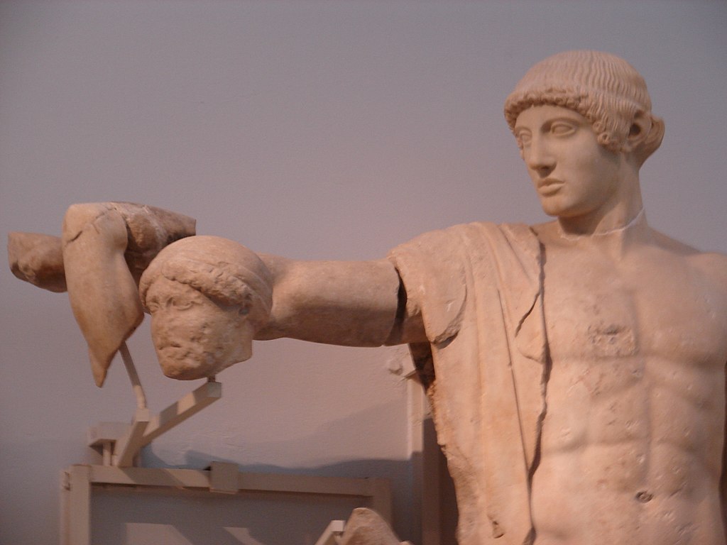 The head and torso of Apollo, a young man. He is nude except for drapings over one shoulder, and his hair is short and curly. Beside him on a stand are arm and head fragments of a statue of Peirithos, a bearded man.