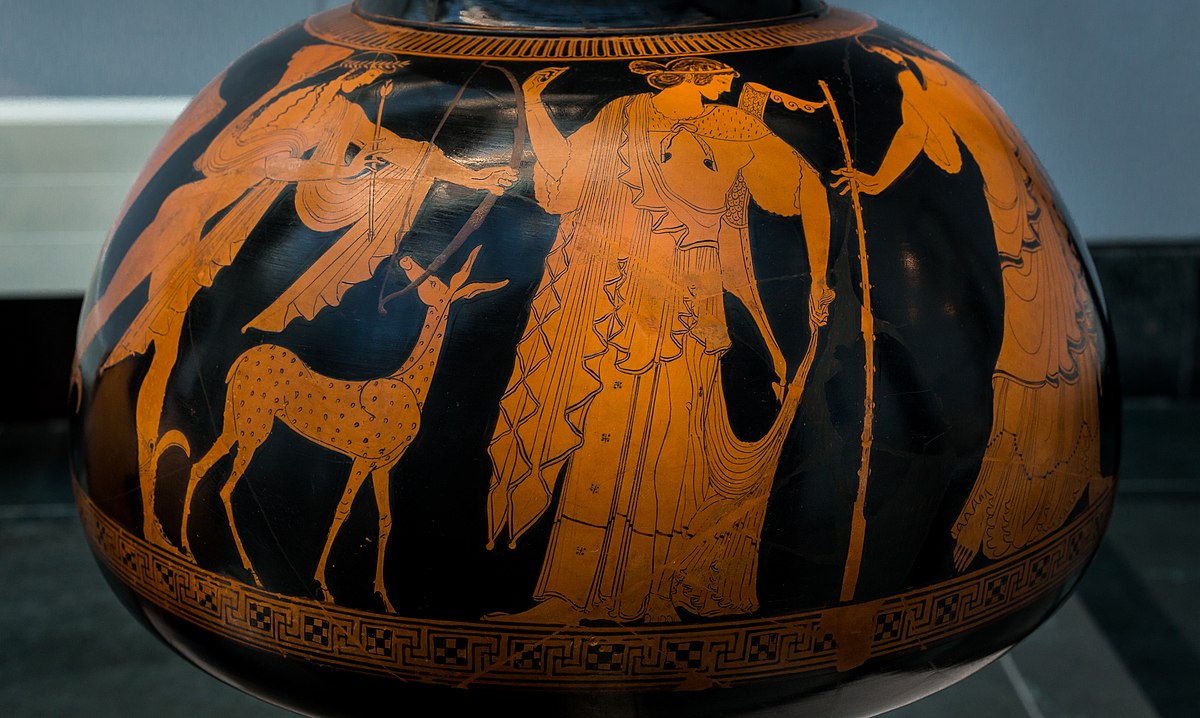 Artemis, draped in an elaborate Himation and with a bow on her back. She looks over her shoulder at a bearded man who stands behind her. To her left is Apollo, shooting a bow, and a deer.