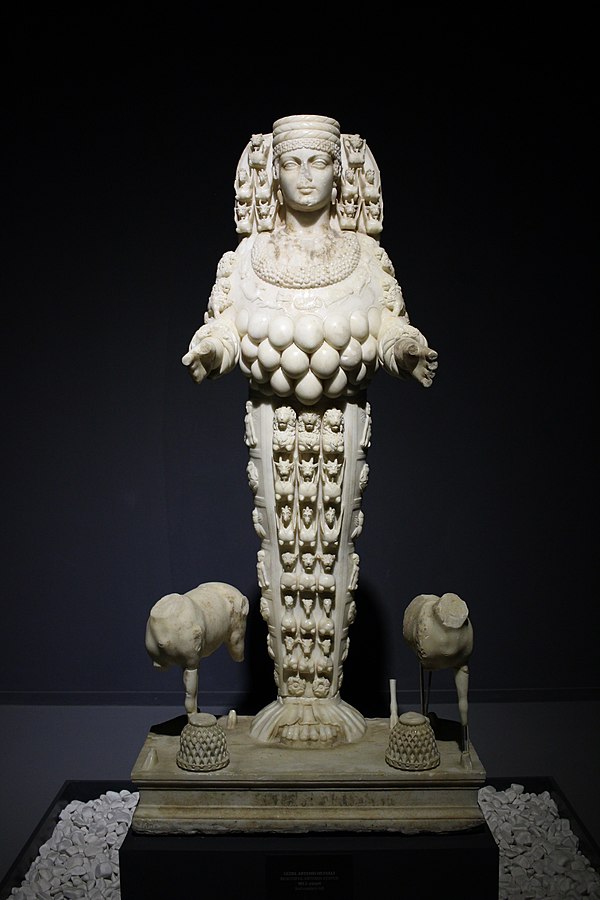 Artemis stands looking straight ahead, her expression placid. Her skirt is elaborately carved with the heads of animals. Around her torso hang many pear-shaped protrusions, and on her head is a cylindrical hat. Small dog-like creatures stand to either side of her.