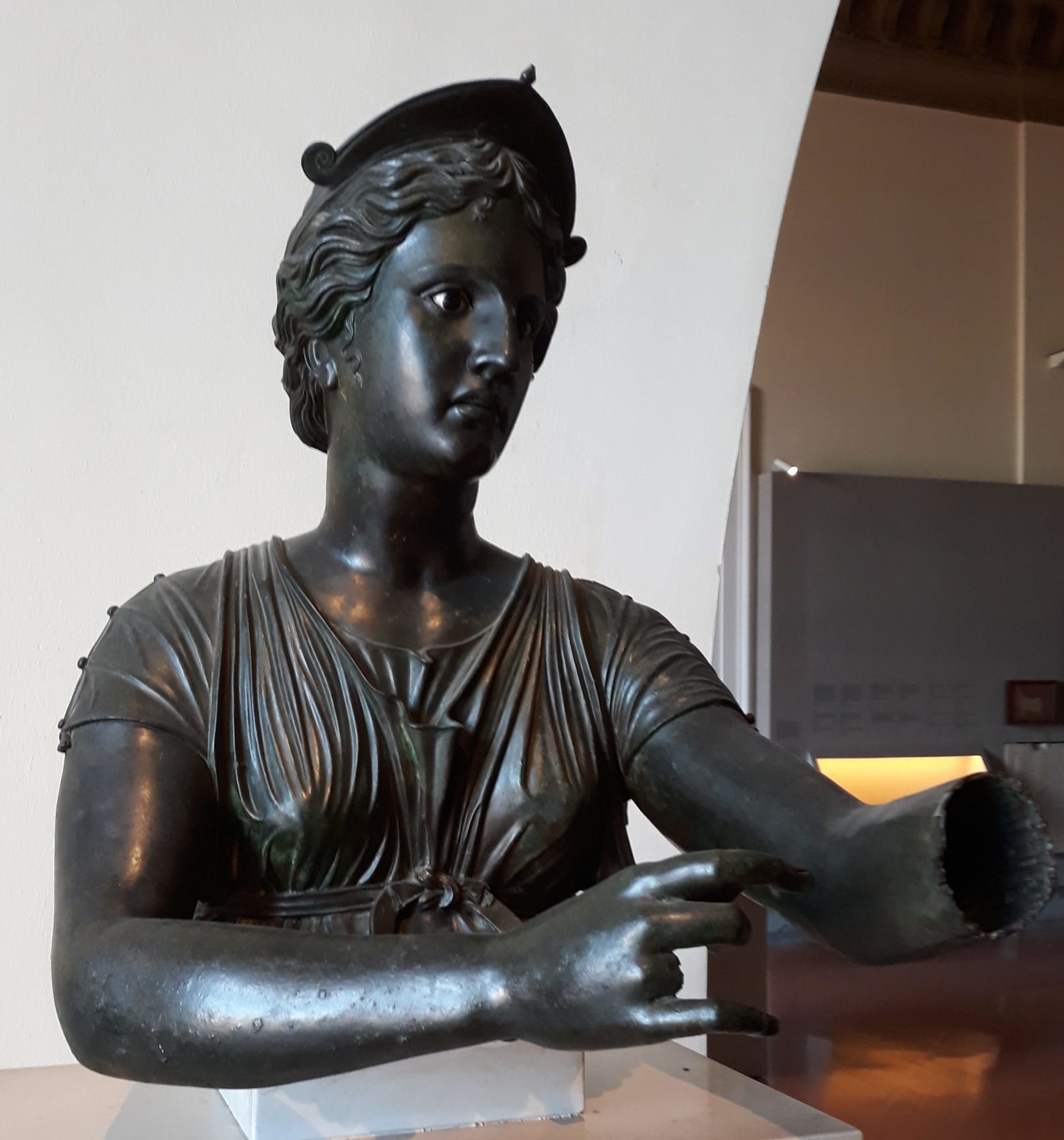 Sculpture of dark bronze of the head and shoulders of Artemis. She has her arms held out in front of her as if she was holding a bow. She wears a crown and is dressed in simple sleeveless garb.