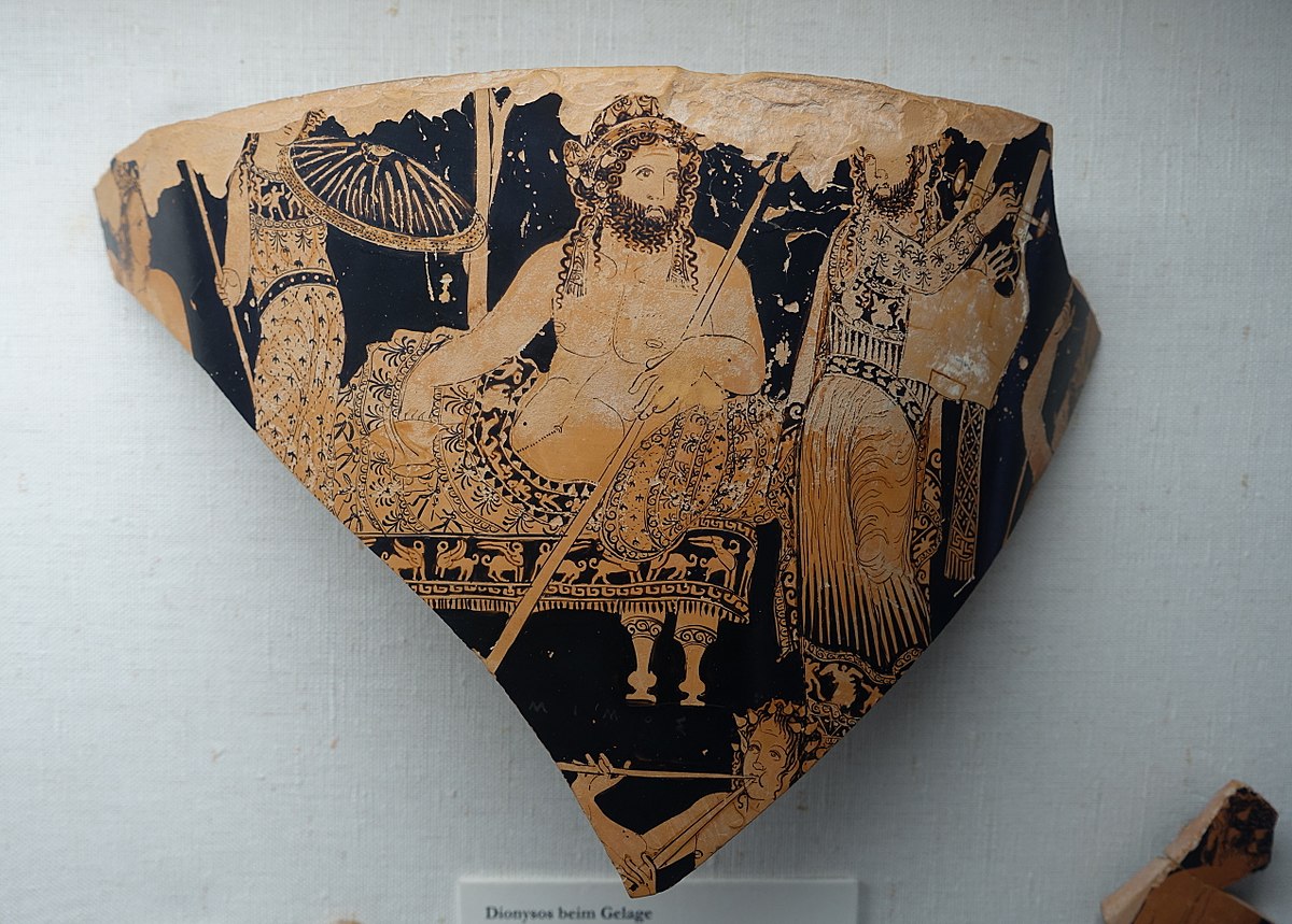 Dionysus reclines on a couch. He has an ornate cloth wrapped around his waiste and wears an elaborate headdress. In one hand he holds a thyrsos, and in the other a cup. Three musicians stand around him.