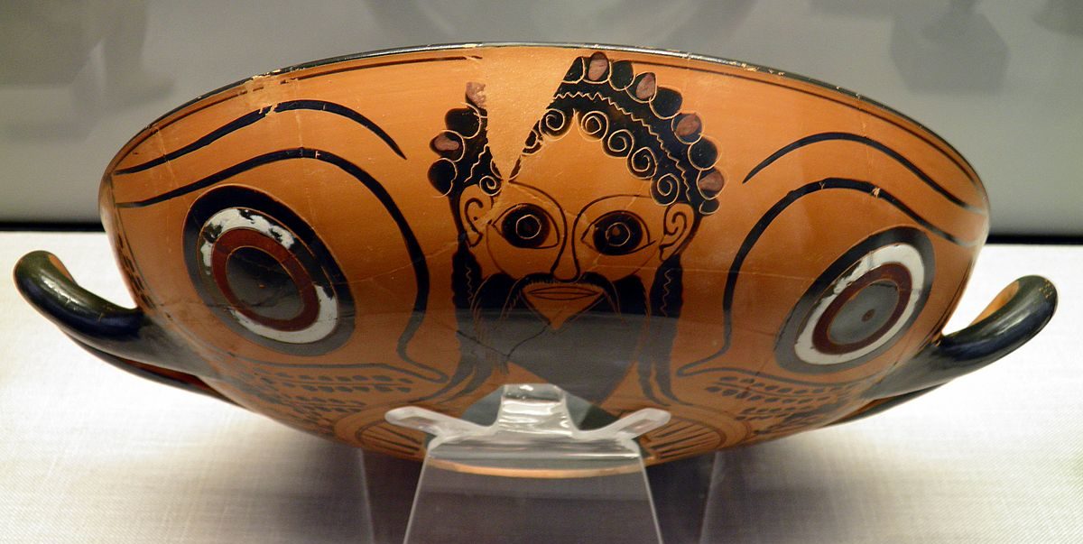 A cup with two large eyes on the outside of the bowl giving the appearance of a face. Between the eyes is the head of Dionysus, bearded and wearing a crown.
