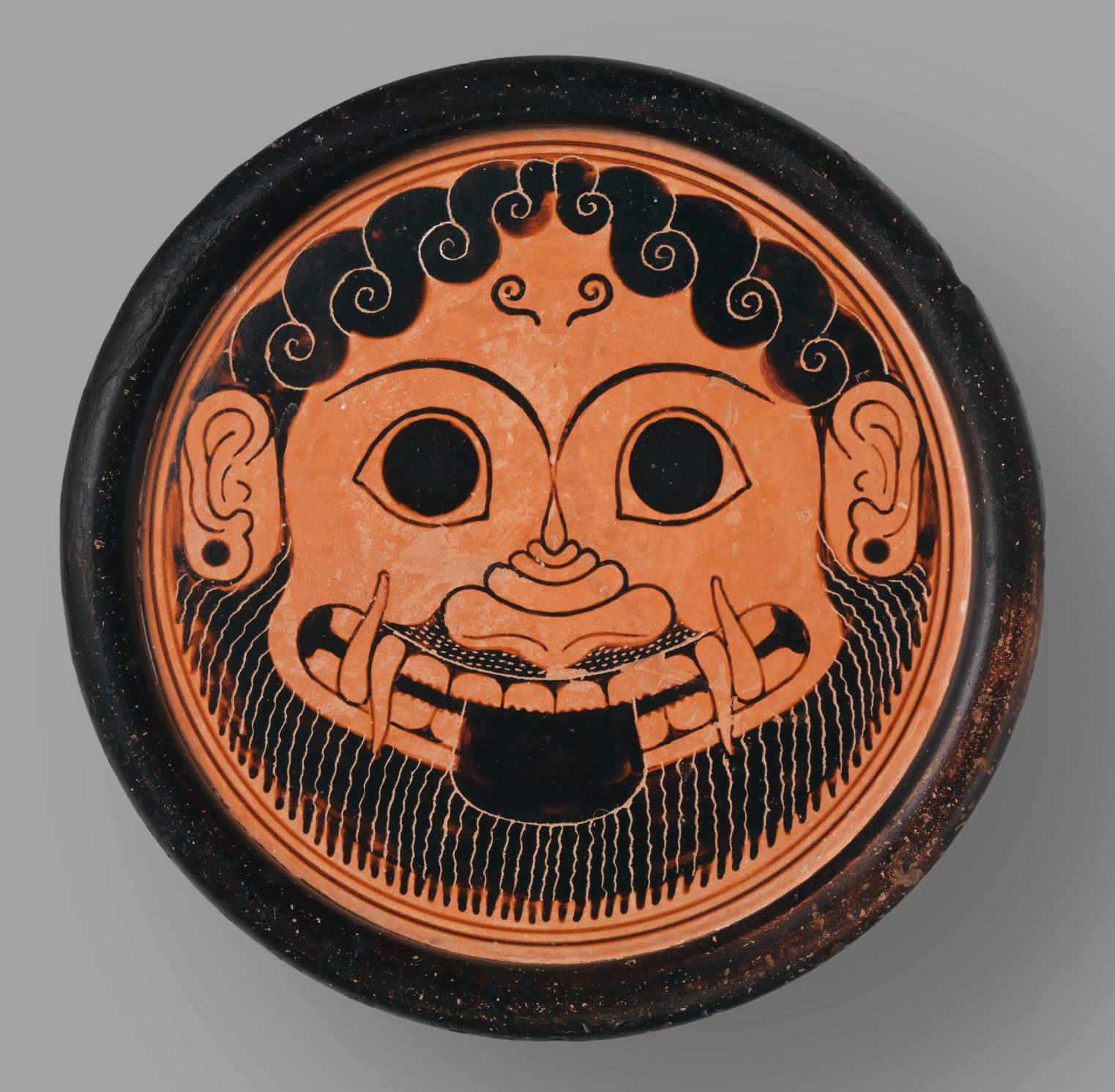 Head of a broad-faced grinning woman. Her smile reveals tusks and her tongue sticks out. She has curly hair and a beard, and wears earrings..