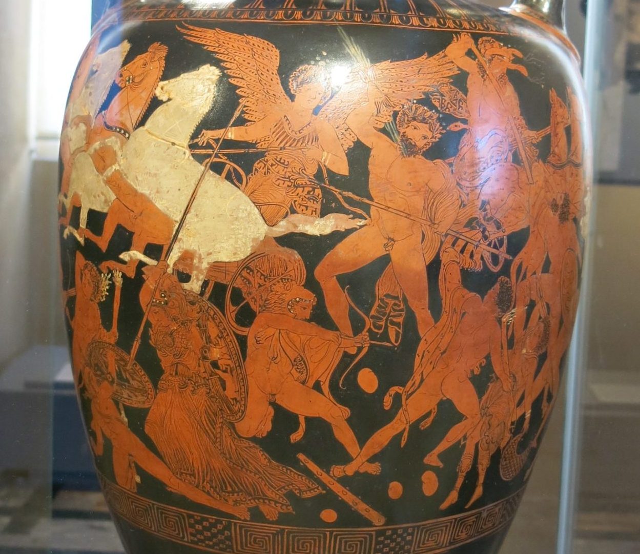 Winged Nike rides in a horse-drawn chariot. Other gods, including Zeus with his lightning bolt, Athena with helm and spear, and Heracles with a bow and lion skin, are engaged in battle with giants.