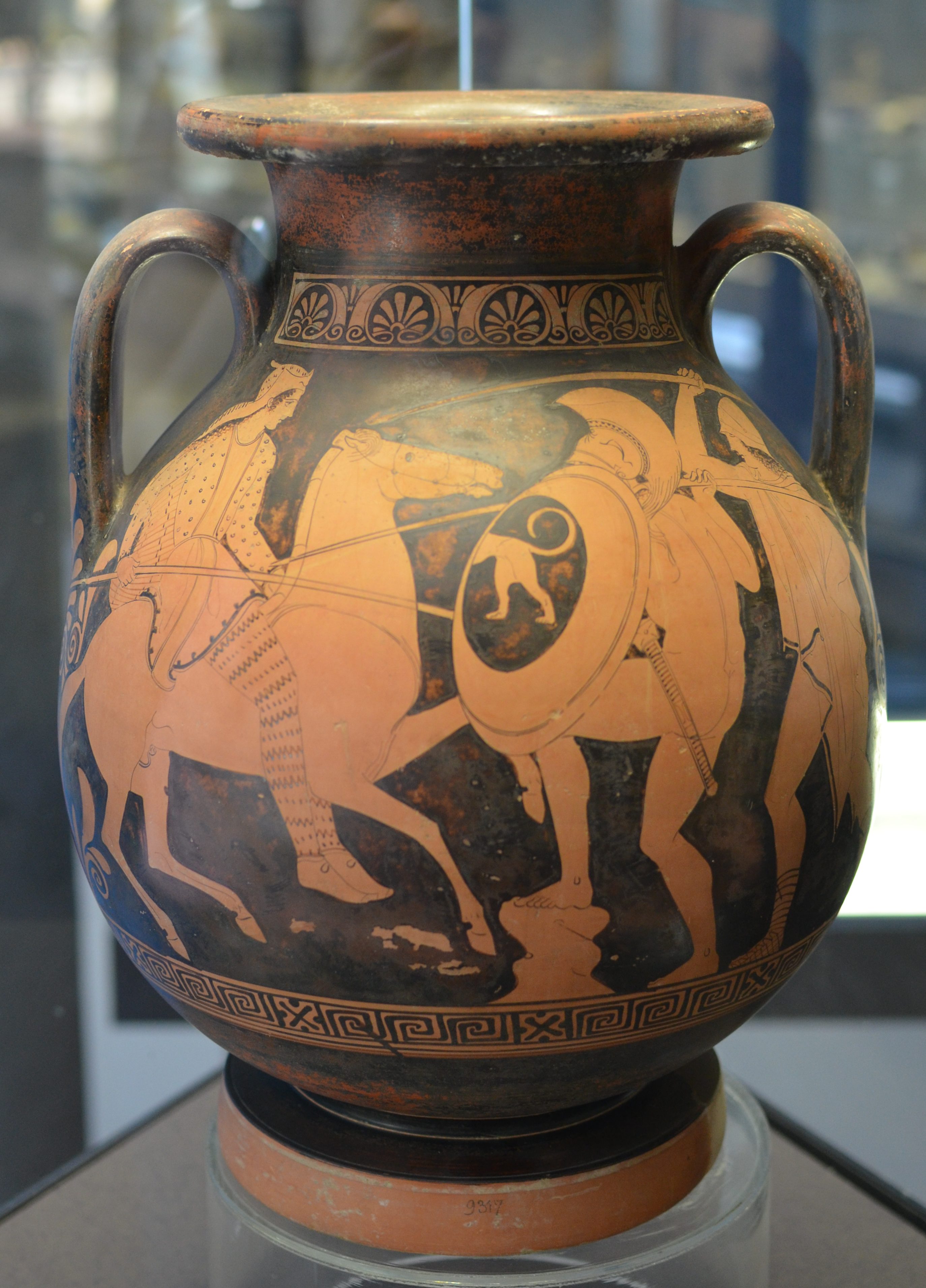 An Amazon, riding a horse, fight a Greek warrior. The Amazon has striped leggings and a hat, and wields a spear. The Greek wield a round shield decorated with a lion, and a spear, and wears a helm.