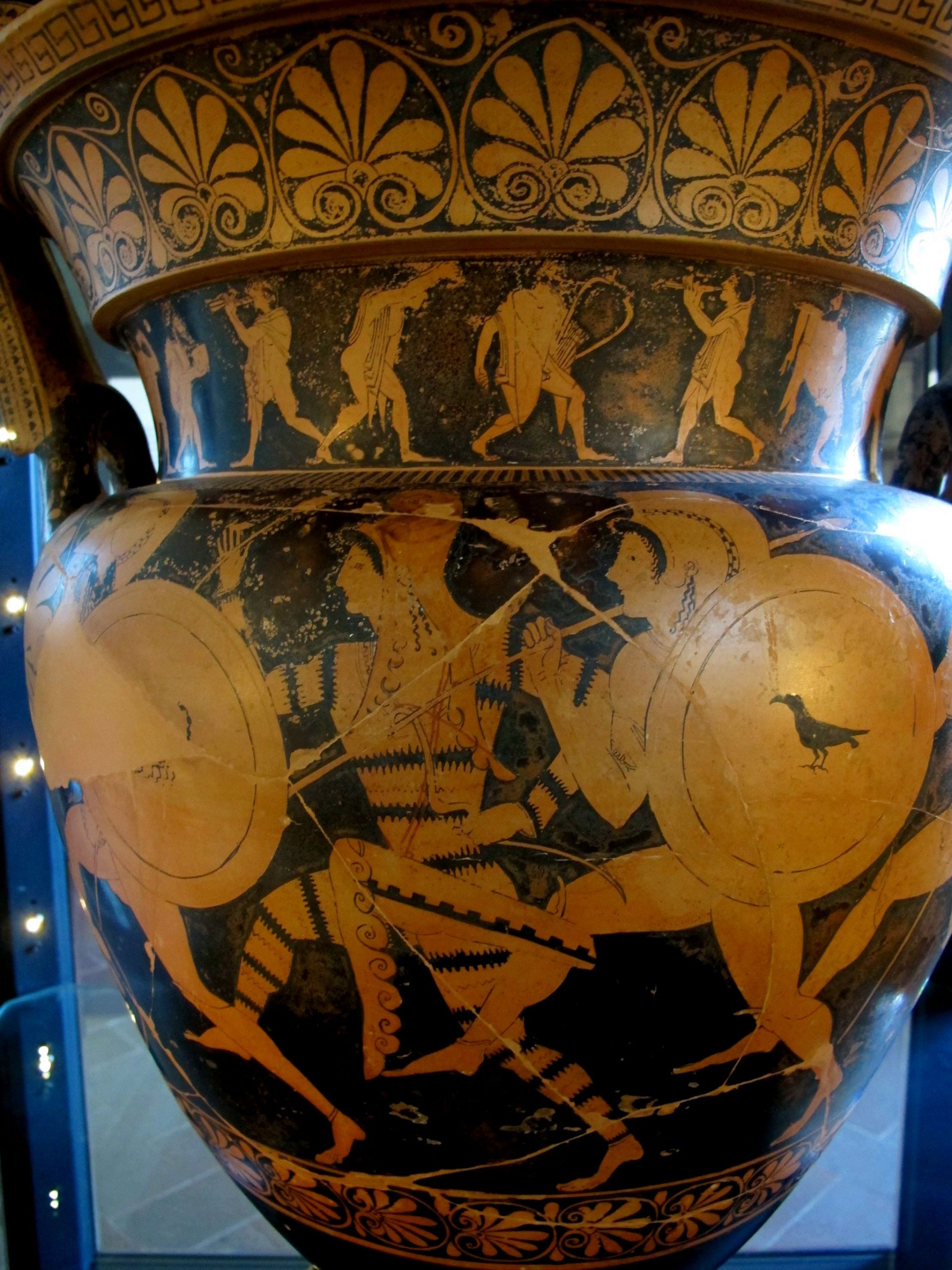 Three figures running in procession. The centre figure is an Amazon, wearing a phrygian cap and tiger-stripe body suit. The other two are Greek warriors, nude with helms and shields.