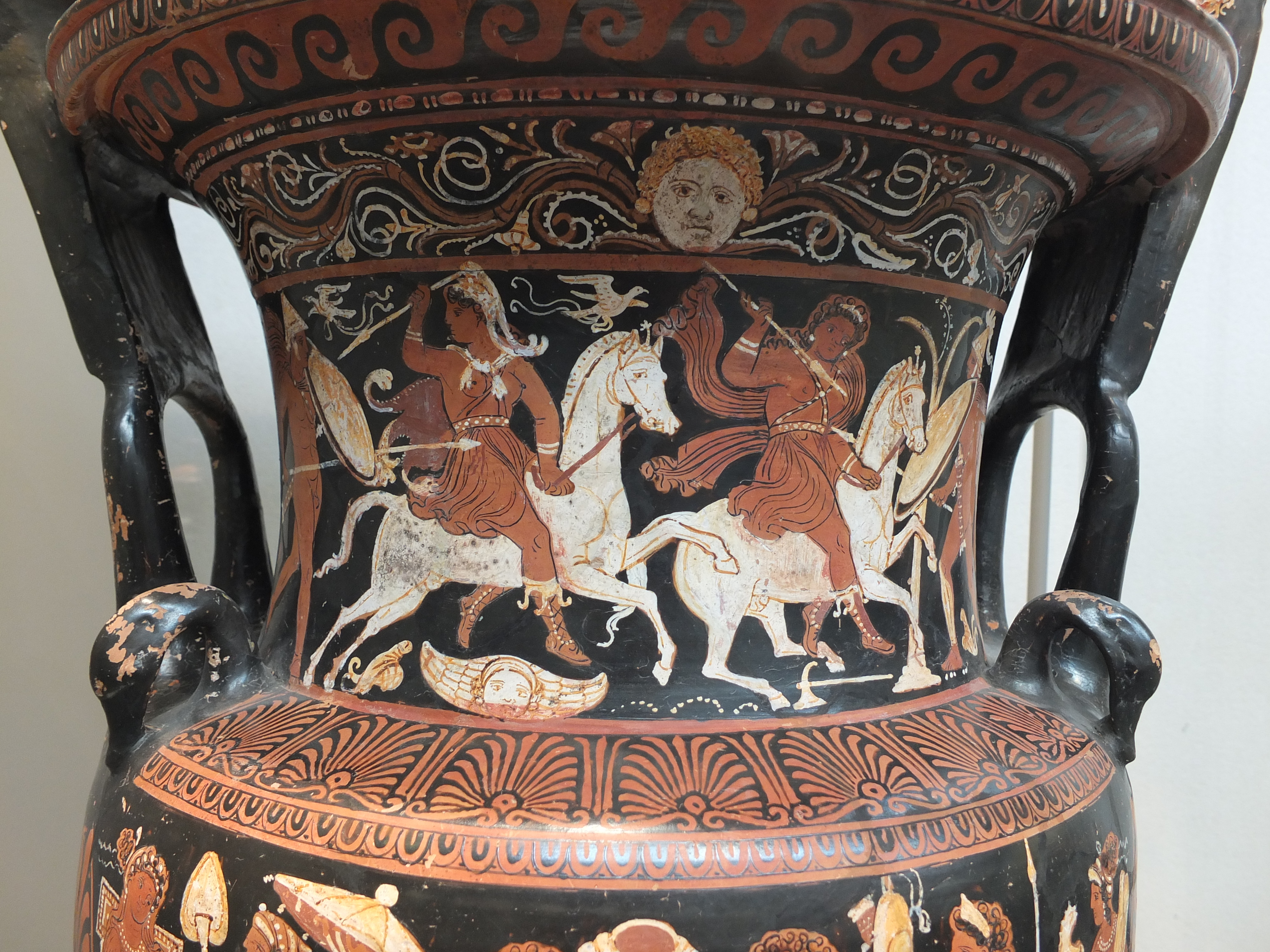 Two Amazons on horses. They have bare chests and wield spears. One wears a hat of wrapped cloth. Birds fly around them. They fight two figures who stand off around the sides of the krater..