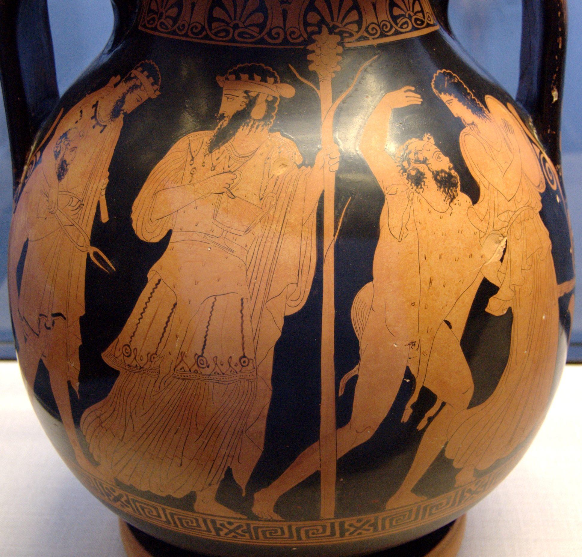 Dionysus, holding his thyrsus, walks ahead of Hephaestus. Hephaestus is slumped and leaning on a man. A silenus and maenad woman walk ahead play instrument.