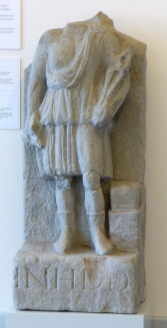 Vulcan in a tunic and holding a pair of tongs. The head of the relief is missing.