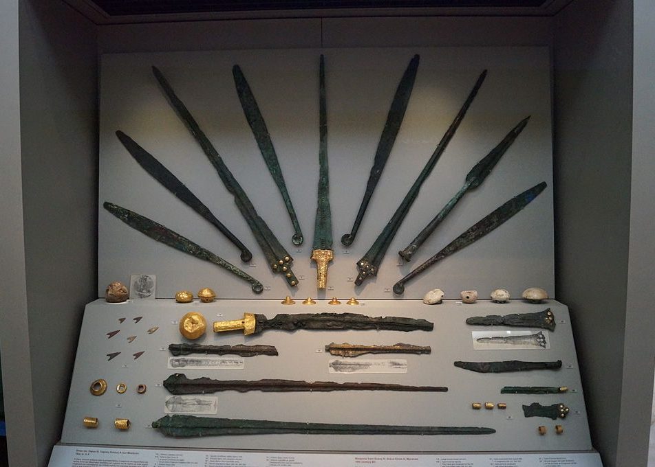 9 dark metal swords are arranged in a fan pattern on the wall. Below, fragments of about 8 more blades, and a collection of arrowheads and small metal rings are displayed.