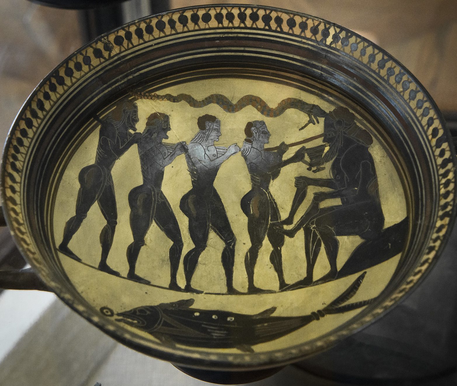 Odysseus and three of his crew, all nude men with long hair. Odysseus holds a stick and holds out a pitcher of wine of Polyphemus. Polyphemus is seated and holds two human legs as he drinks the wine. Odysseus' crew carry a large stick between them. A snake is above the scene, and a large fish below.