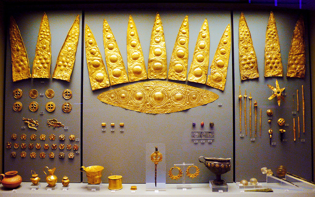 A wall display of gold jewelry. Centre: A diadem with crown-like peaks, decorated with circle patterns. 3 more crown-like diadem pieces are mounted on each side. Below, a collection of brooches and buttons. On the table, a pair of earrings, 7 small vessels, and decorative knives.