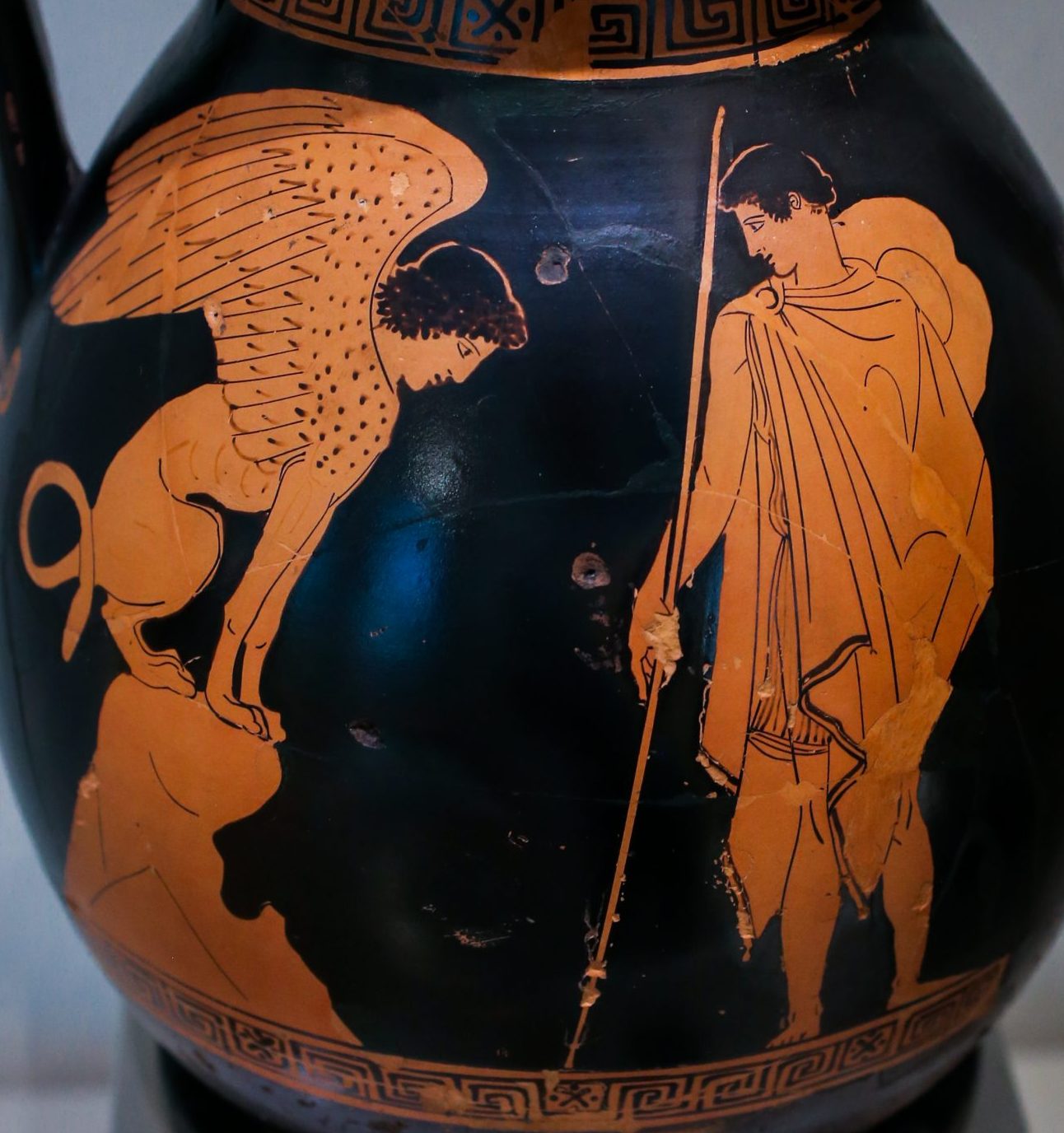 Oedipus, with a chlamys cape and a petasos hat around his neck, stands holding a long sceptre. The sphinx, a winged lion with a humanoid head, sits on a rock in front of Oedipus.