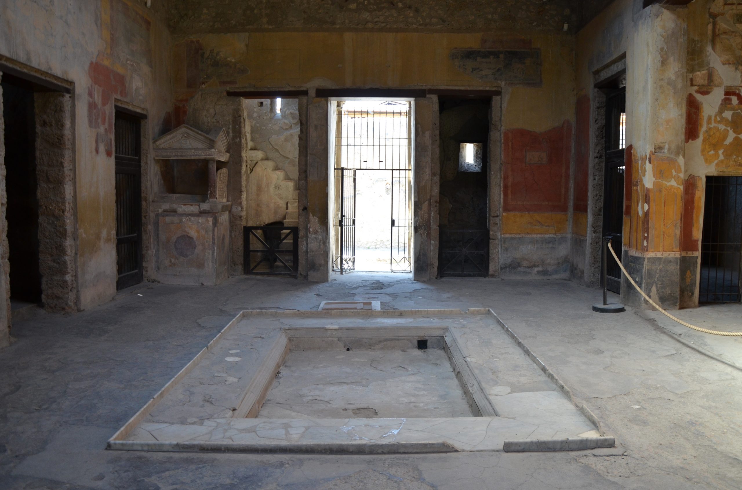Atrium. In the far corner, a small temple-style lararium set against the wall. The centre of the floor has rectangular shallow pit.