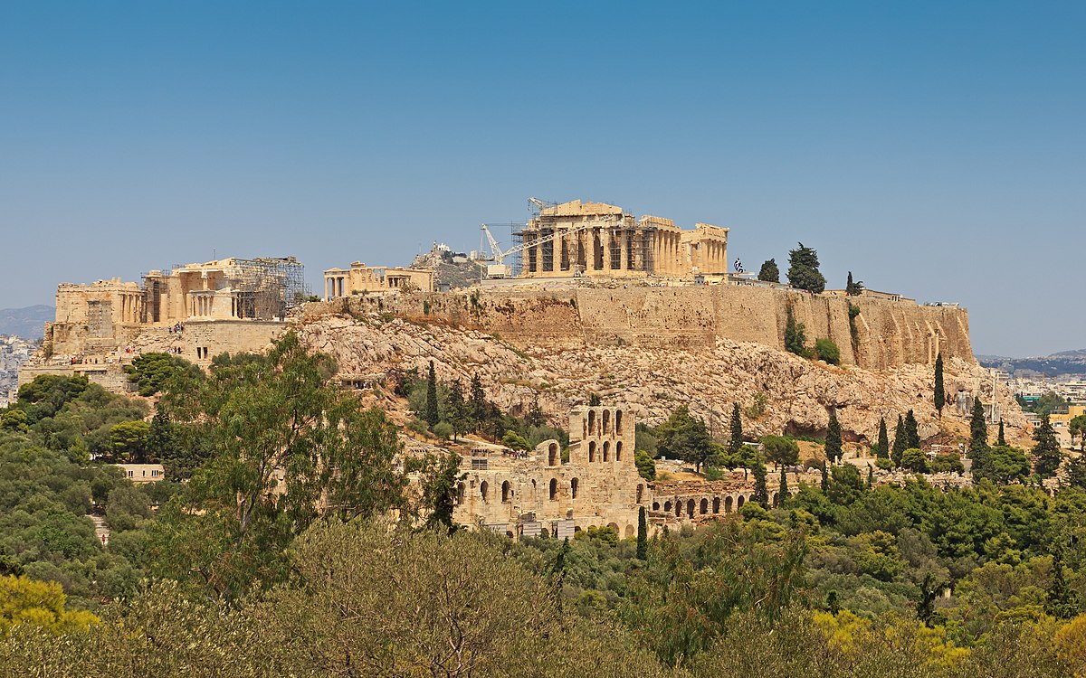 Archaeological remains of Athens: the Parthenon and other buildings are on top of a hill, the Acropolis. At the food of the hill, more buildings are visible amongst trees.