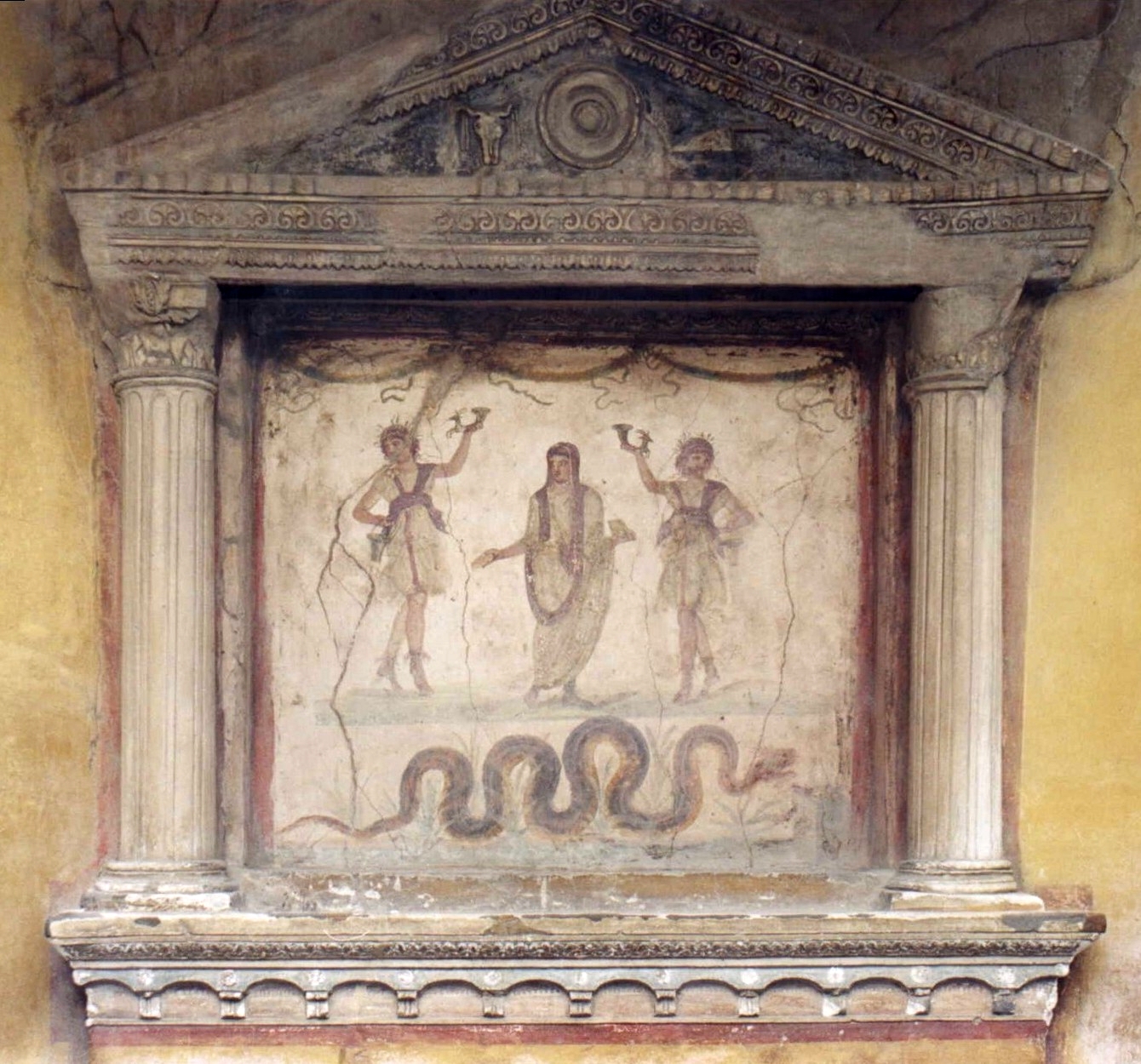 A lararium shrine framed by temple-like columns and pediment. Inside the columns, a fresco depicting the genius in a purple-trimmed toga, flanked by two lares holding cornucopias. A large snake in grass slithers beneath their feet.