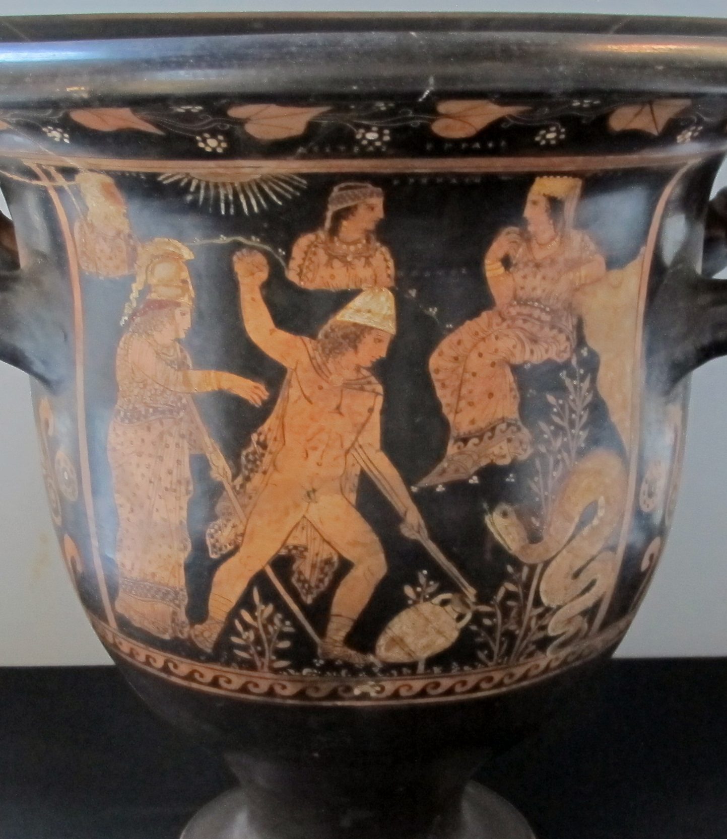 Cadmus, nude with a chlamys cape and a rounded helm. He has one fist raised, and an amphora lies on the ground by his other handA snake-like dragon is coiled by a tree in front of him. Three figures sit in the sky above Cadmus, and Athena stands behind him.