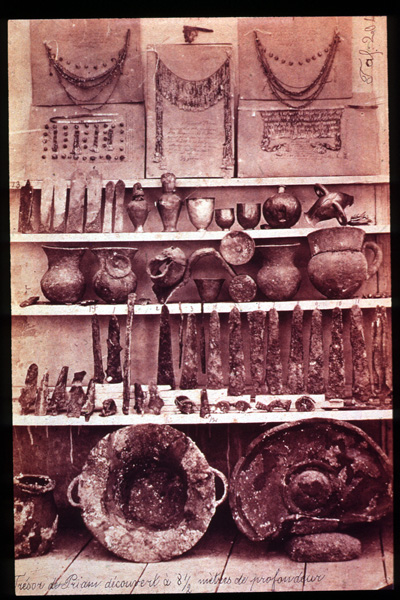 Four display shelves of jewelry, vessels, and blades. Top shelf: three large necklaces. 2nd and 3rd shelves: a collection of metal vessels and pots. 4th shelf: a collection of metal blade fragments. Floor: A large metal pot and a circular shield.